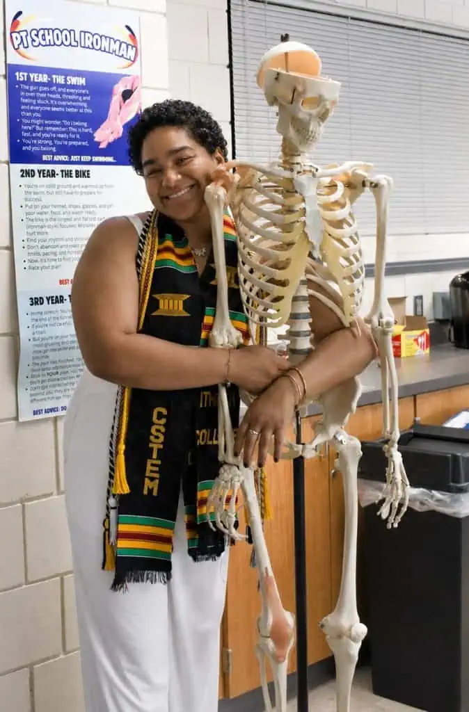 Anika, wearing their kente graduation stole, laughs while hugging a teaching skeleton in the physical therapy lab.