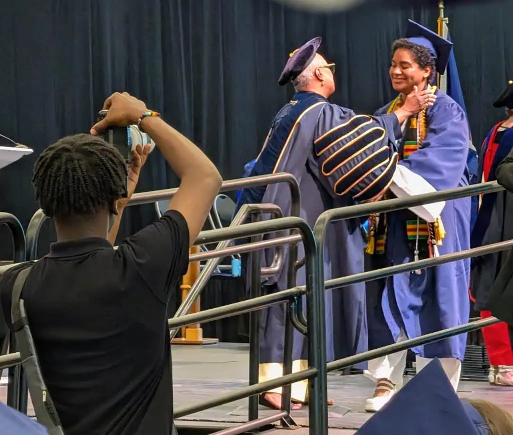 Anika, a young biracial Black graduate in a navy cap and gown with a kente stole, smiles as they cross the stage and the Ithaca College president, a Black woman in academic robes and a tam, opens her arms to draw them into a hug. In the foreground, a young person raises a phone to capture the moment.