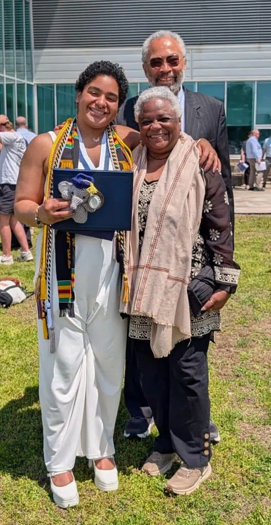 Anika in graduation clothes stands with Gwen and Roger, Black elders and chosen family who drove from Ohio. Roger stands behind, Gwen beside Anika.
