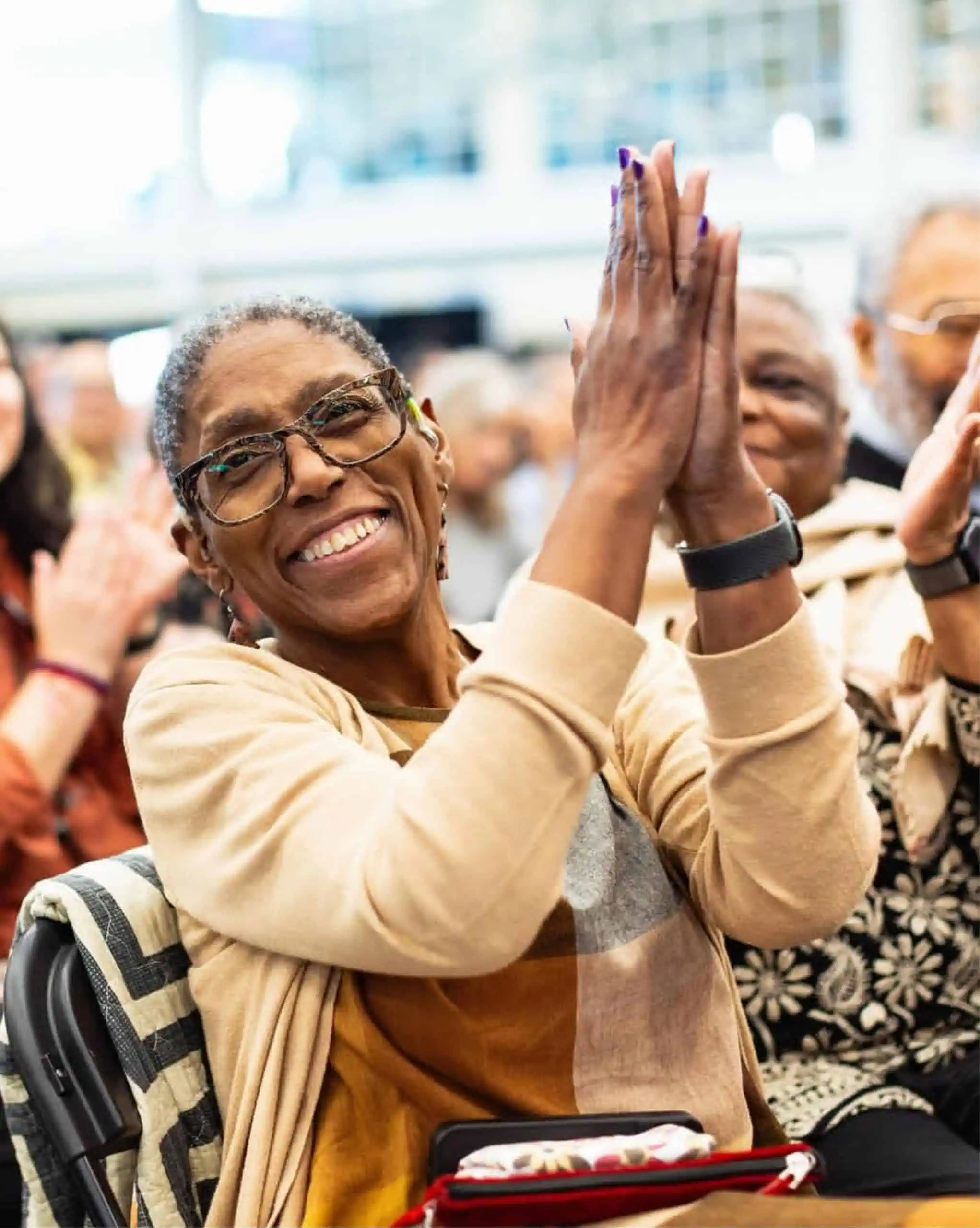 Roxy Manning, a Black woman in glasses, beams and claps with raised hands, caught mid-cheer as she watches her child Anika graduate.